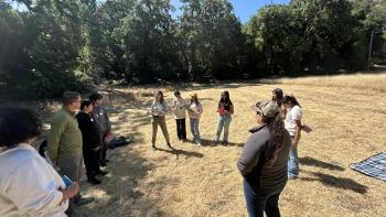 A group of people are gathered in a circle outdoors on a field covered in dry, light brownish-tan grass, and a large number of deciduous trees surround the field.