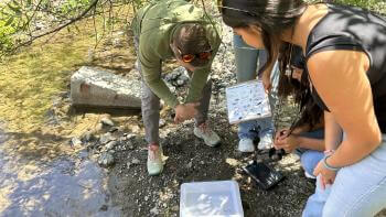 Students and an adult gathered near a stream, examining aquatic samples. One holds a clipboard with charts, another uses a magnifying tool.