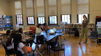 A woman, standing and gesturing, leads a discussion with attentive students seated around tables. The classroom has large windows and bookshelves, conveying an educational atmosphere.