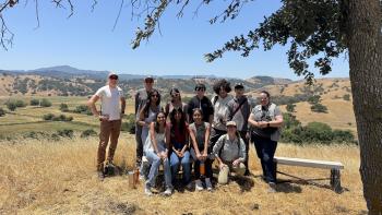 A group of ten people stands and sits on a grassy hill overlooking a valley. A large oak tree provides shade for the group.