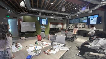 A large conference room with students gathered around several tables, engaged in discussion. An adult stands in front of a whiteboard labeled "Social Media Promos."