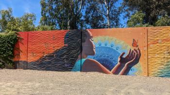 A large colorful mural on an outdoor wall showing a girl with long dark hair reaching out her hands to a small monarch butterfly, in front of a orange background and blue mandala design