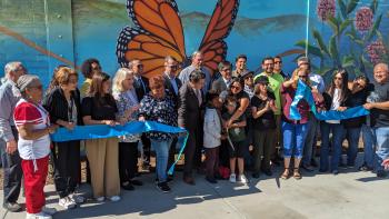 A large group of adults and children stand in front of a large colorful mural with a monarch butterfly, behind a recently cut blue ceremonial ribbon