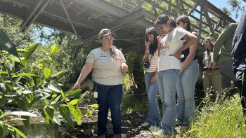 A group of young people learn about a native plant from an adult while standing under a bridge.