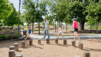 A family of four stands on individual wooden logs that are laid out in a semicircle at a park.