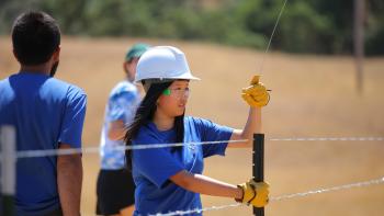 A young person in a hard hat attaches a wire to a fence post.