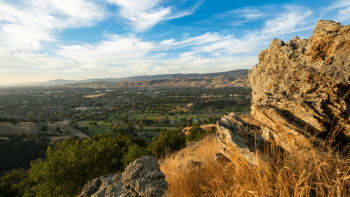 A rocky cliff overlooks a valley below with clouds above and a blue sky.