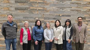 Seven board of directors standing in front of gray wood paneled wall