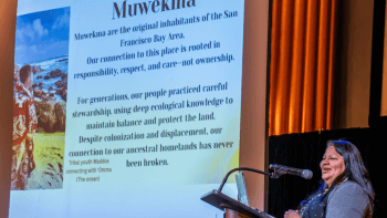 Bernadette Quiroz stands at a podium with a screen behind her that shares information about the Muwekma