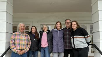 Group of six adults standing on a covered porch smiling at the camera