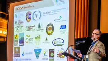 A man stands at a podium talking into a microphone next to a screen with a projection that reads "SCMSN Members" with various logos beneath it. 