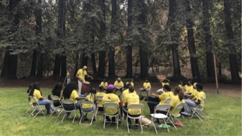 A group of people sit in a circle of chairs on grass, surrounded by redwood trees.