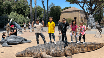 Five children standing on large, life-sized crocodile statue in a sunny park with trees, people, and a stone building in the background.