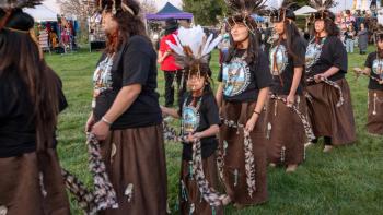 Group of members from Muwekma Ohlone Tribe in traditional dress with feathered headdresses walking in a cultural festival procession outdoors.