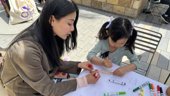 A mother and her daughter sit at a table and color a wooden snake with markers.