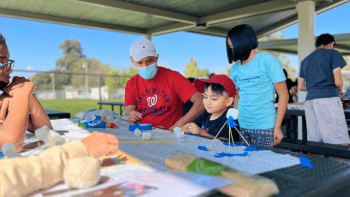 A boy in a red hat sitting at a covered picnic table with two adults working on a craft activity