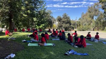 A group of people wearing red t-shirts sitting on yoga mats in a shady grass area under trees