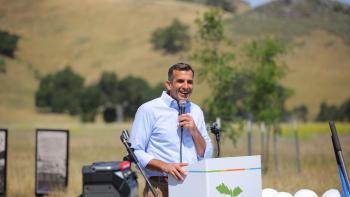 Congressman Sam Liccardo stands at a podium with a microphone in hand with trees and a golden hill in the background.