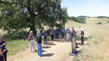 A group of people gather in a circle on a nature trail to look at a mock up of the future interpretive signage that will be on display in this location.