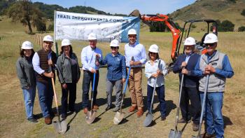 Elected Officials stand in front of a nature trail, construction equipment and a sign that says "Trail Improvements Coming Soon" while wearing hard hats and holding shovels.