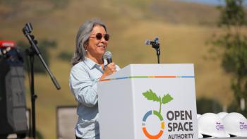 Director Kathy Sutherland stands at a podium with a microphone in hand addressing an audience with a golden hill in the background.