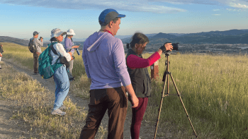 A woman positions her camera on a tripod to take a photo with the help of a man as they stand in a grassy path surrounded by nature