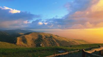 A photo of a sunset breaking through fluffy clouds taken from the top of a mountain with the view of another green mountain in the distance.