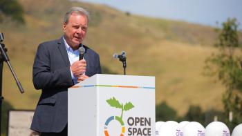 Senator Dave Cortese stands at a podium with a microphone in hand and a golden hill in the background.