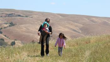 A mom and two small children walk through the grass on a hill with rolling golden hills in the background.