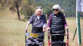 Two women with mobility devices stand on the path of a nature trail.