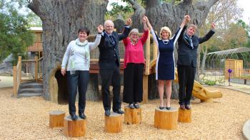  Five people stand on wooden stumps in a playground, holding hands and raising their free arms in celebration.