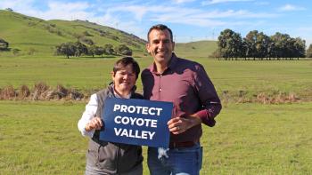 Sam Liccardo and Andrea Mackenzie stand in a sunny, grassy field, holding a blue sign that reads "PROTECT COYOTE VALLEY," and smiling. Behind them are rolling green hills, scattered trees, and a clear blue sky with wispy clouds.