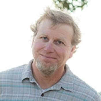 light skinned man with white goatee wearing a light blue patterned shirt against a white background