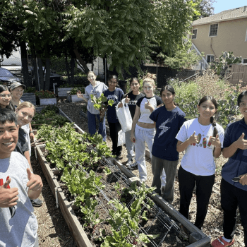 garden box in the center of 2 rows of young people with their thumbs up with trees in the background and a 2 story home on the right side