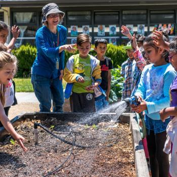 A group of young students in colorful clothing stand smiling and laughing around a raised garden bed, one of the students is holding a sprinkler towards the soil in the bed; a teacher stands to one corner of the bed pointing and smiling at the students