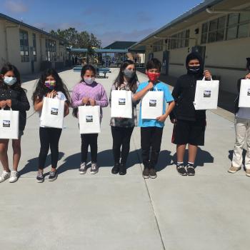 A row of seven children standing outside of a classroom area holding up white paper bags with educational supplies