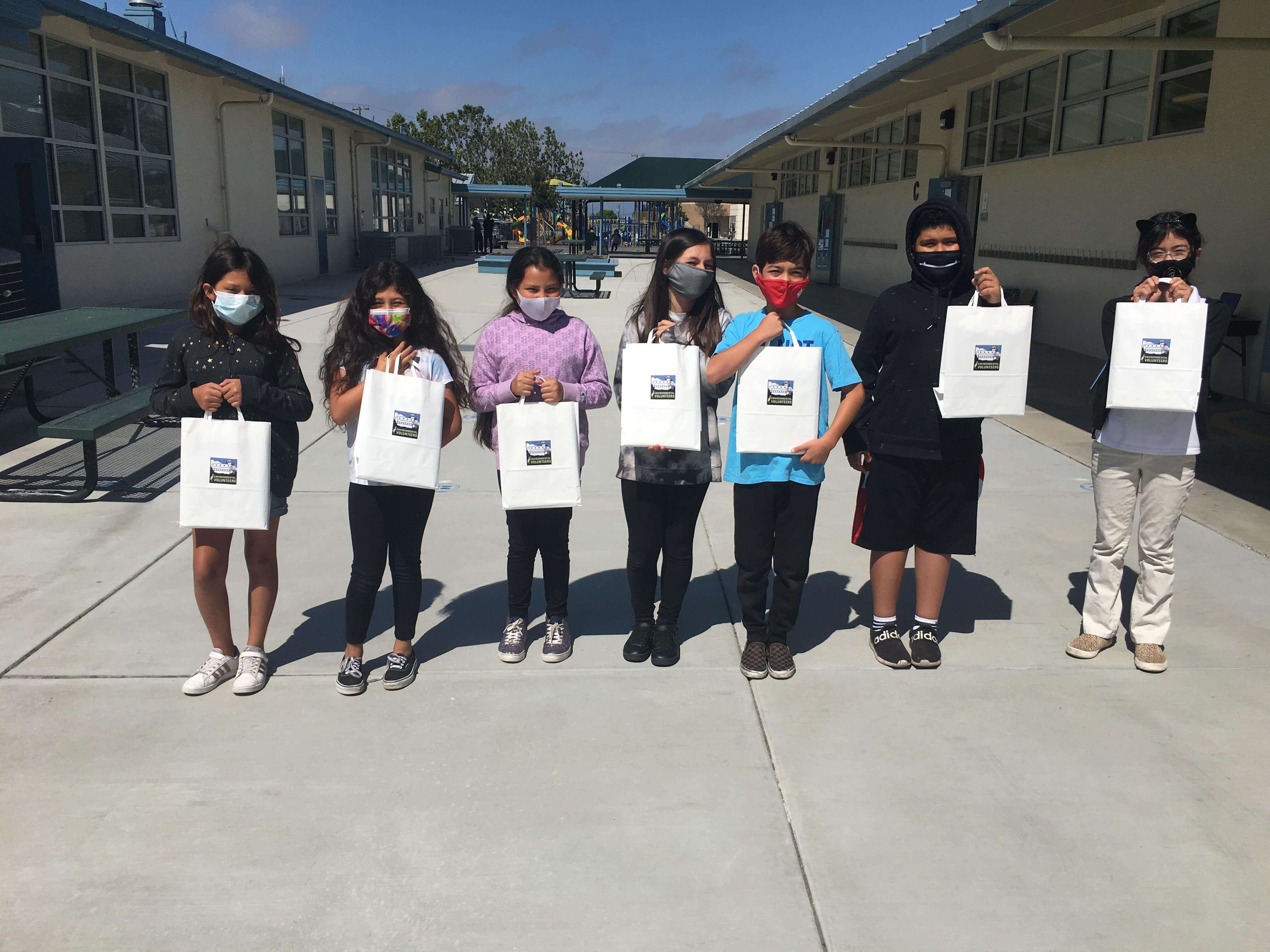 A row of seven children standing outside of a classroom area holding up white paper bags with educational supplies