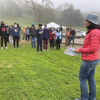 A woman in a red jacket stands in the foreground facing a group of people wearing rain coats standing on a grassy field with a mist-covered hillside in the background