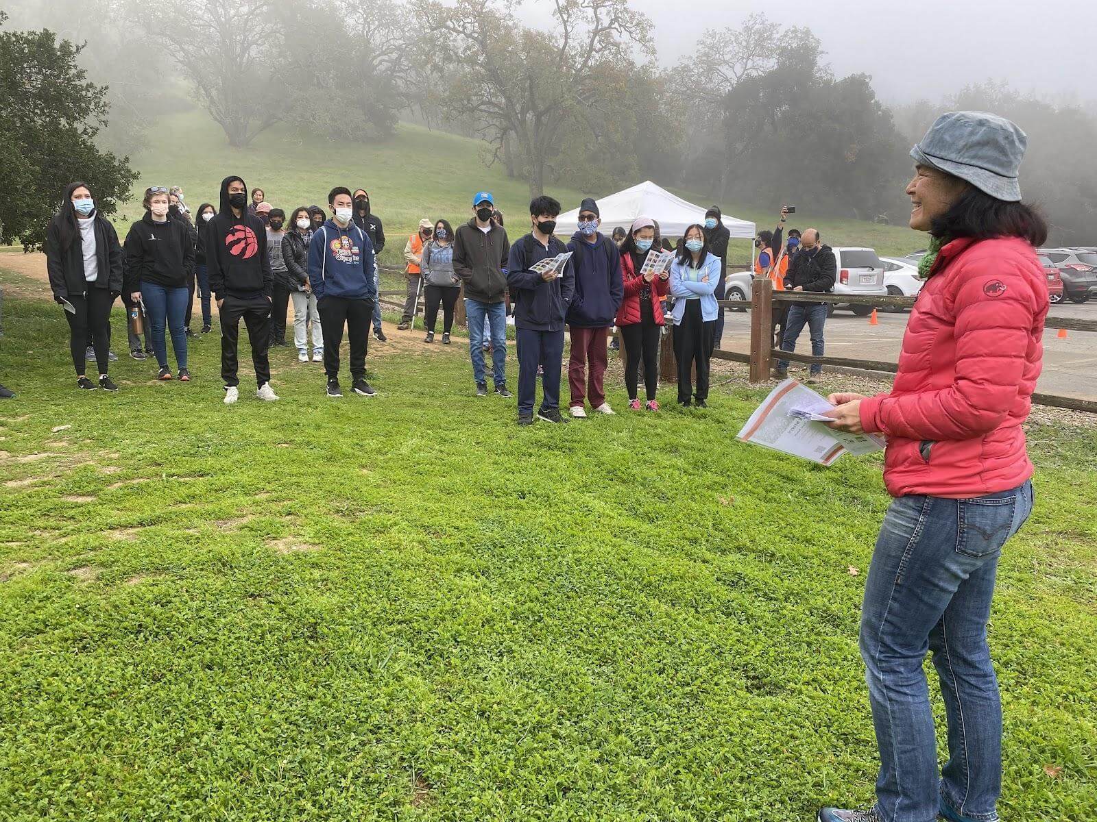 A woman in a red jacket stands in the foreground facing a group of people wearing rain coats standing on a grassy field with a mist-covered hillside in the background
