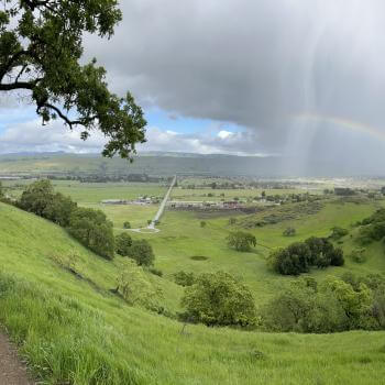 A dirt trail winds through lush green hills dotted with dark green oak trees, in the distance a rainbow arcs over farmland below