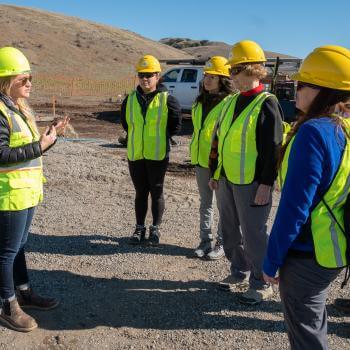 Blonde woman in yellow hard hat and vest speaking to a group also wearing yellow hard hats and vets against a dirt hill an light blue sky