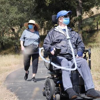 A person wearing a blue face mask in a motorized wheelchair is followed by a person also wearing a blue face mask walking on a paved nature trail. 