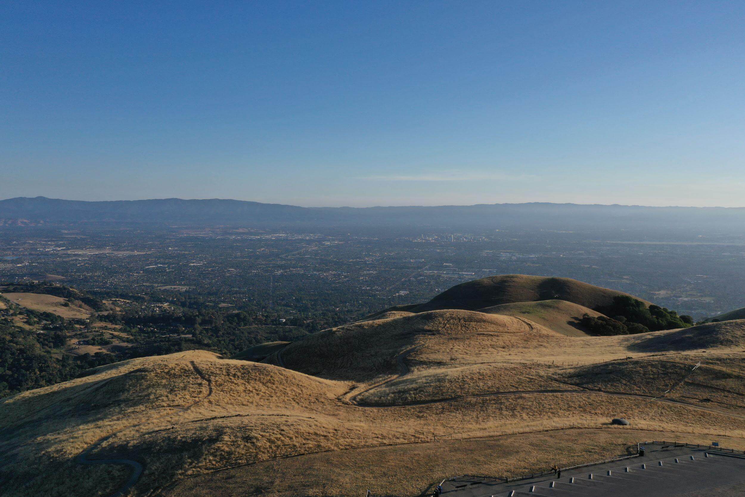 An aerial view of Sierra Vista's rolling gold hillsides with trails winding across them. Beyond the hills is an expansive view of Santa Clara Valley below, under a clear blue sky.