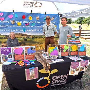Three people stand smiling at a camera under a booth and behind a table with the Open Space Authority logo and covered in colorful brochures and informational items about Coyote Valley