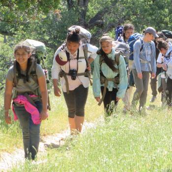 A line of girls wearing backpacking backpacks walk along a trail bordered by tall grass, smiling at the camera