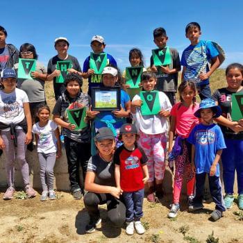 A group of 17 children stand outside smiling at the camera holding up paper with letters that spell "THANK YOU"