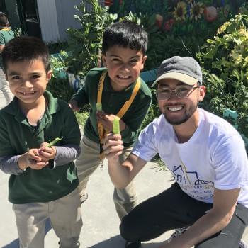 Two young children in green polo shirts stand next to a kneeling young adult in a white t-shirt, holding up pea pods and smiling