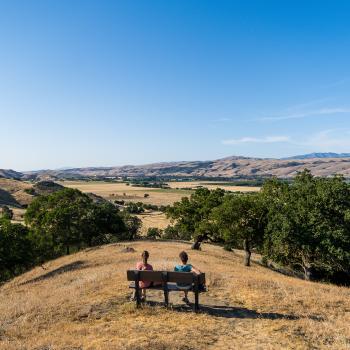 Two hikers sit on a bench overlooking golden hills and trees