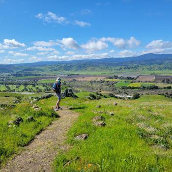 Hiker on a trail on a green grassy field overlooking a beautiful view of a valley patchworked with farm fields and blue mountains in the distance
