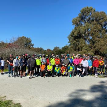 A group of about 50 people wearing bicycle helmets standing on a gravel trail in front of trees and vegetation standing and kneeling while smiling at the camera
